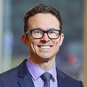 Brian Sinotte wearing a blue suit, glasses and smiling.