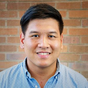 Bert Chantarat smiling while wearing a blue dress shirt in front of a brick wall.