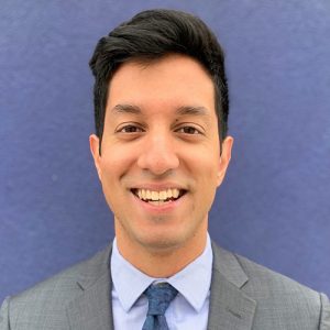 Rohan Khazanchi in a suit and tie standing in front of a blue background.