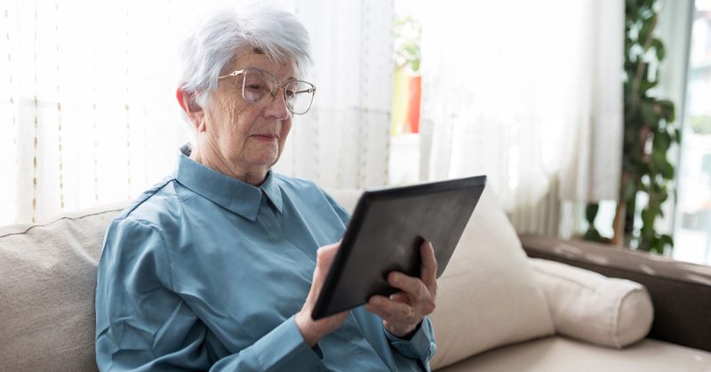 An older woman in a blue shirt using a tablet computer while sitting on a white couch.