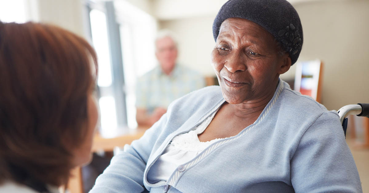 An again Black woman sitting in a wheelchair talking to someone in a nursing home.