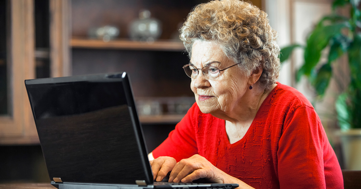 An elderly woman in a red sweater uses a laptop computer.