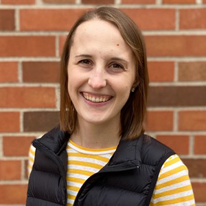 Cecily Weber in a yellow-and-white-striped shirt and black vest smiling in front of a brick wall.