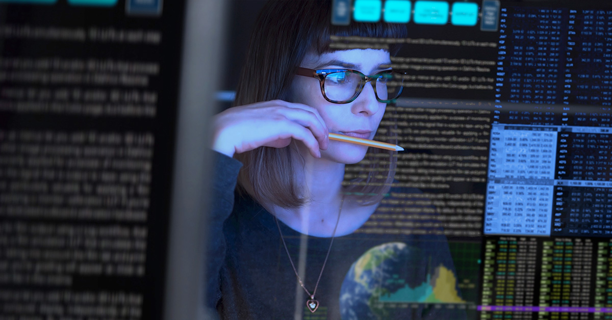 The reflection of a woman's face is seen in computer monitors displaying data and code.