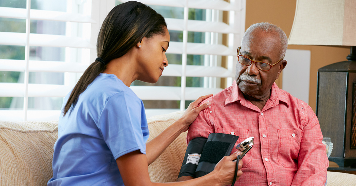 A Black home healthcare worker checks the blood pressure of a Black man sitting on a couch.