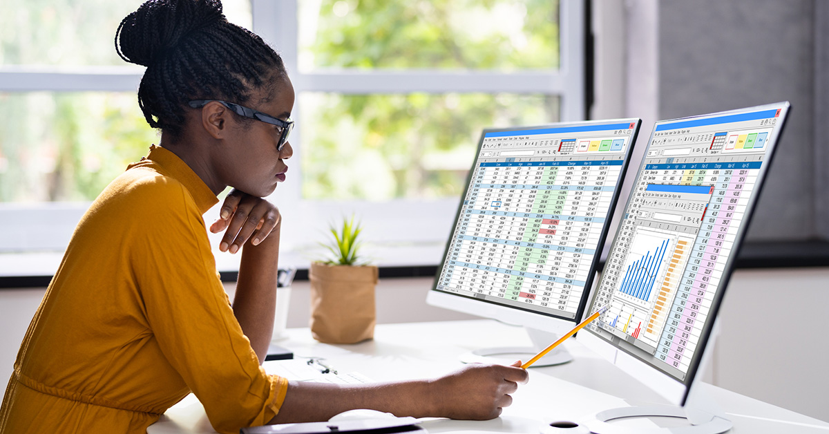 A Black woman wearing a marigold dress reviews data on two computer monitors.