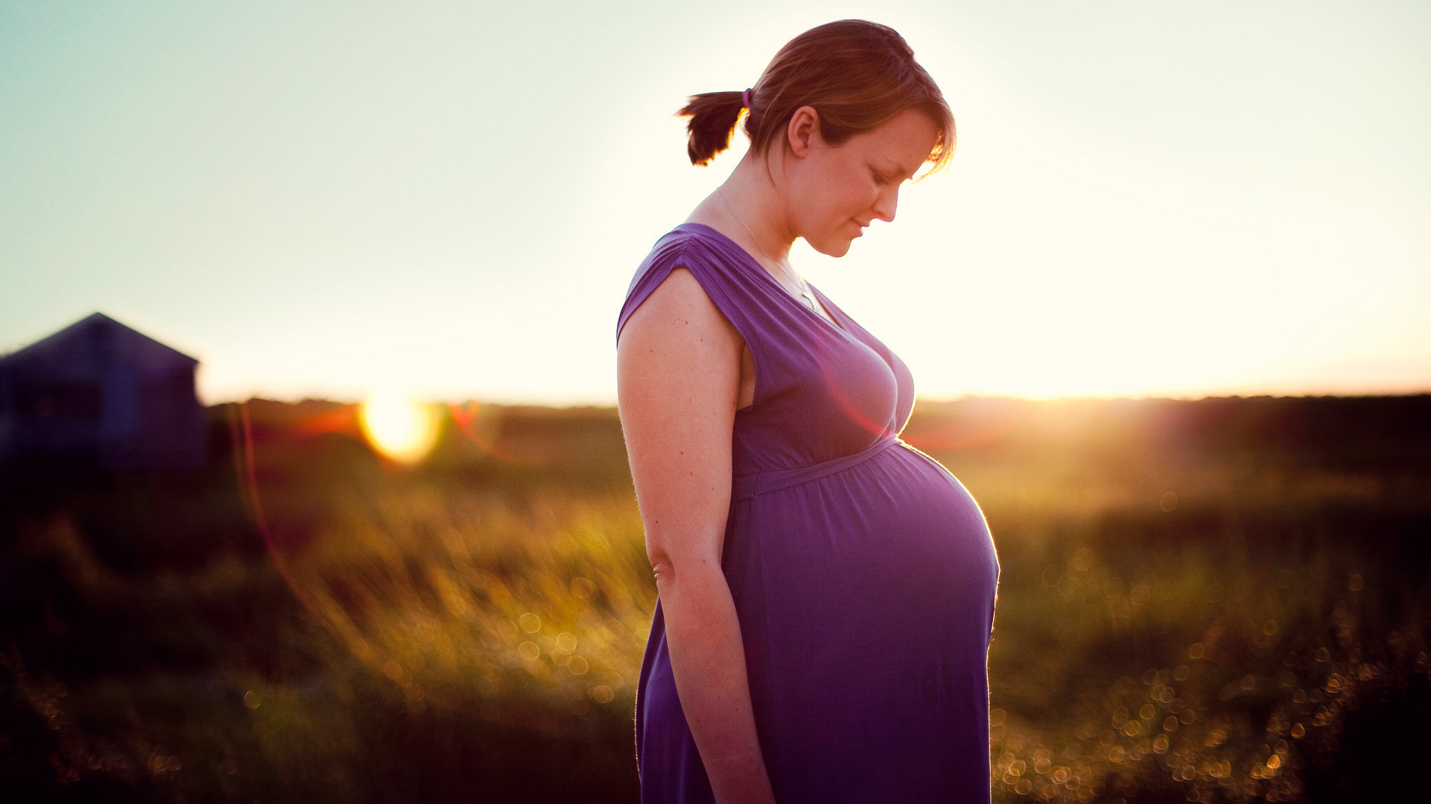 A pregnant woman in a purple dress stands in a field at sunset.