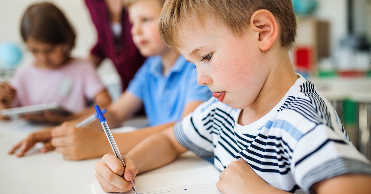 A boy is writing on a page with a pen in a classroom.