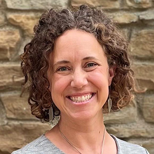 Carrie Henning-Smith smiling in a gray shirt while standing in front of a stone wall.