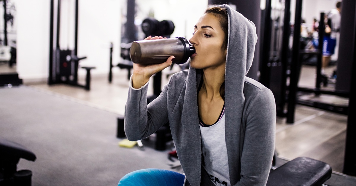 A woman in a gray hoodie drinking a protein shake while sitting on a weight lifting bench.