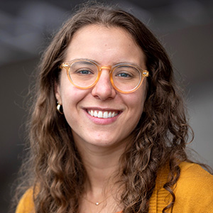 Sarah Samorodnitsky smiling while wearing a yellow shirt in front of a dark background.