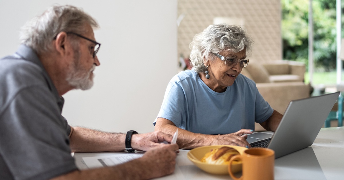 Older man and woman seated at a kitchen table, both looking at a laptop which the woman is typing on.