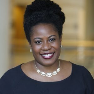 Rachel Hardeman, a smiling Black woman with dark brown skin, brown eyes, and curly natural black hair, wearing a black top with pearl and diamond accessories.