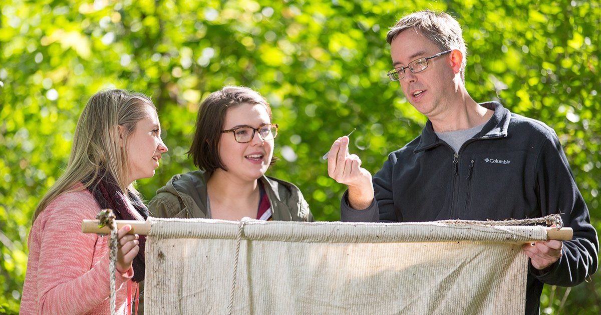 Jon Oliver standing with two women holding a white cloth and examining a tick
