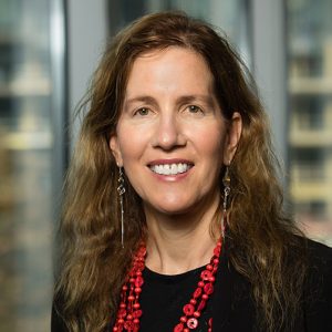 Dianne Neumark-Sztainer smiling while wearing a black shirt and red necklace in front of office windows.