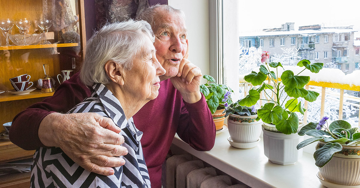 An elderly man and woman cuddle while looking out a window.