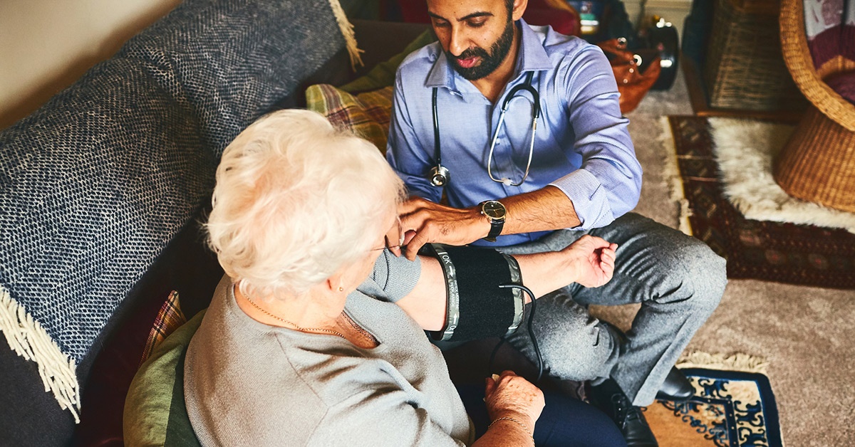 A man measures an older woman's blood pressure in her home.