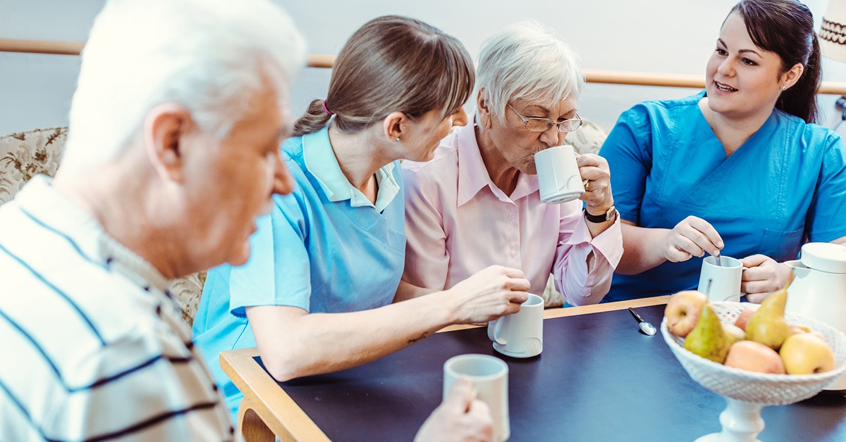 Nursing home residents and staff drinking coffee around a table.