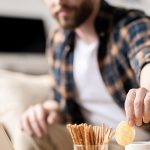 A man grabs a potato chip while working on his laptop from a living room.