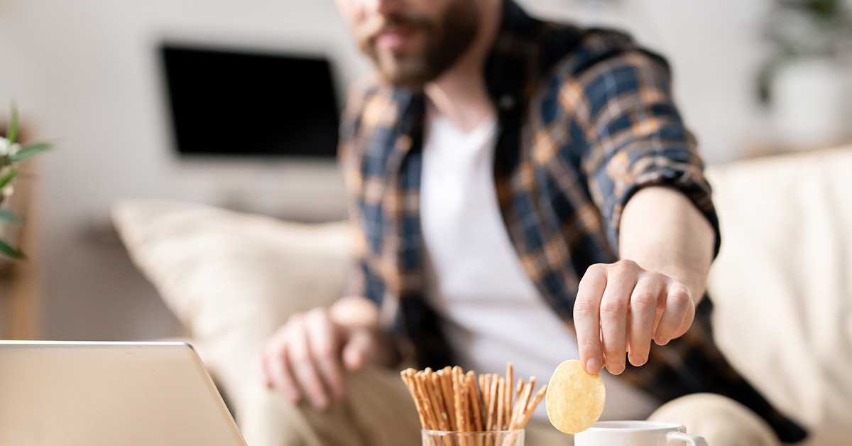 A man grabs a potato chip while working on his laptop from a living room.