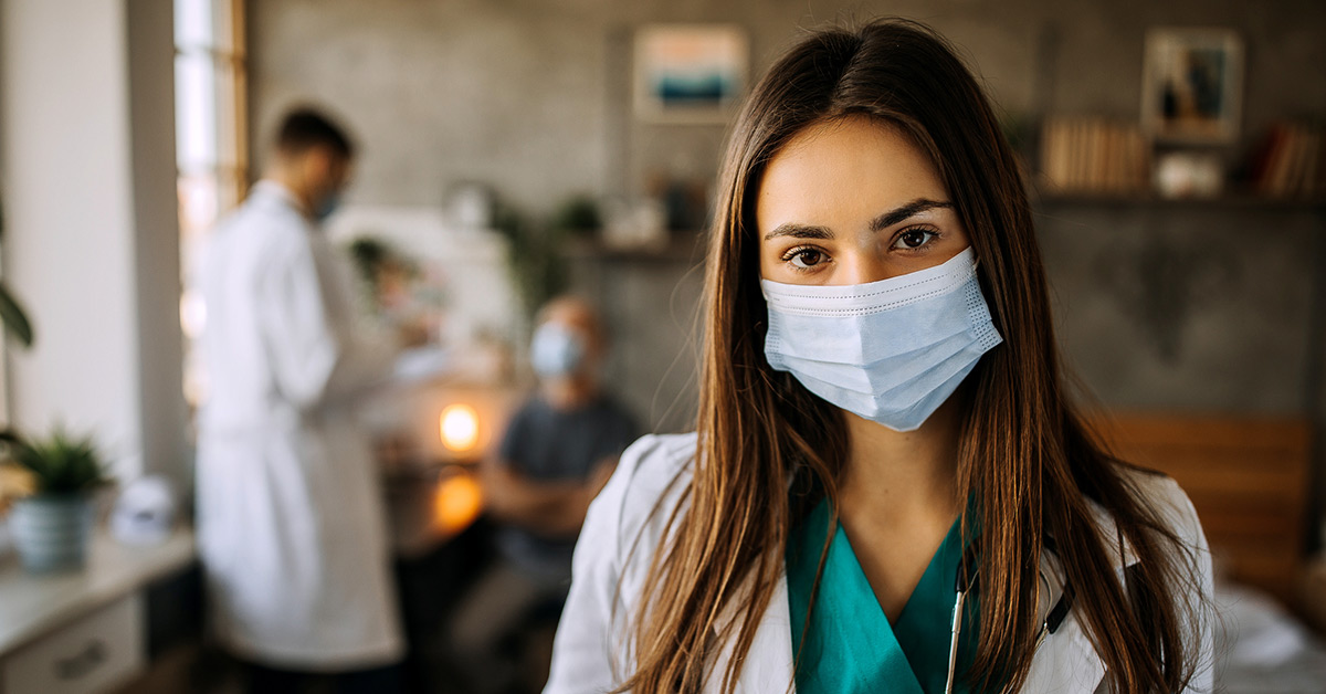 A female medical student wearing a mask, green scrubs, and a white coat.