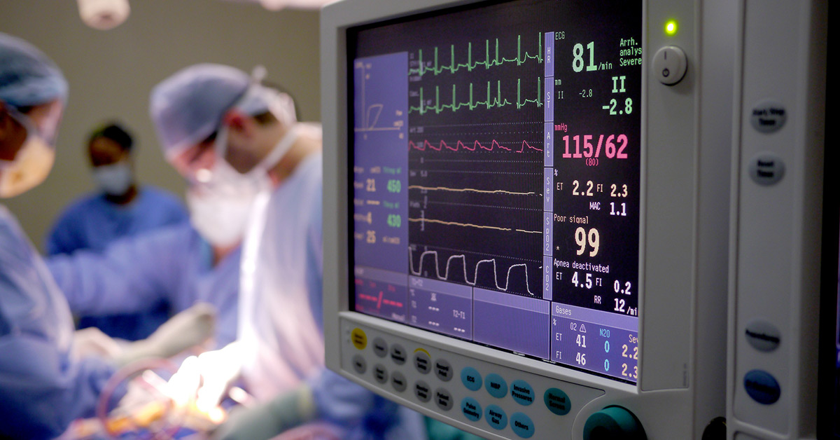 A patient health monitor with a surgery team operating in the background.