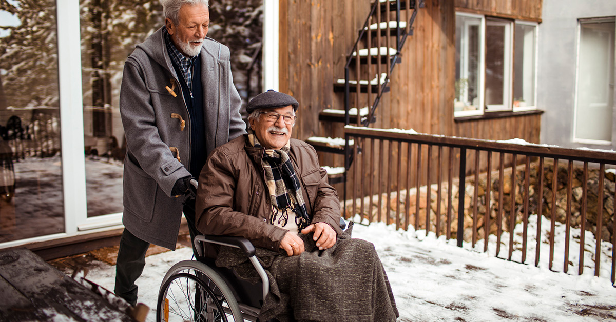An older man wheeling another older man in a wheelchair through the snow.