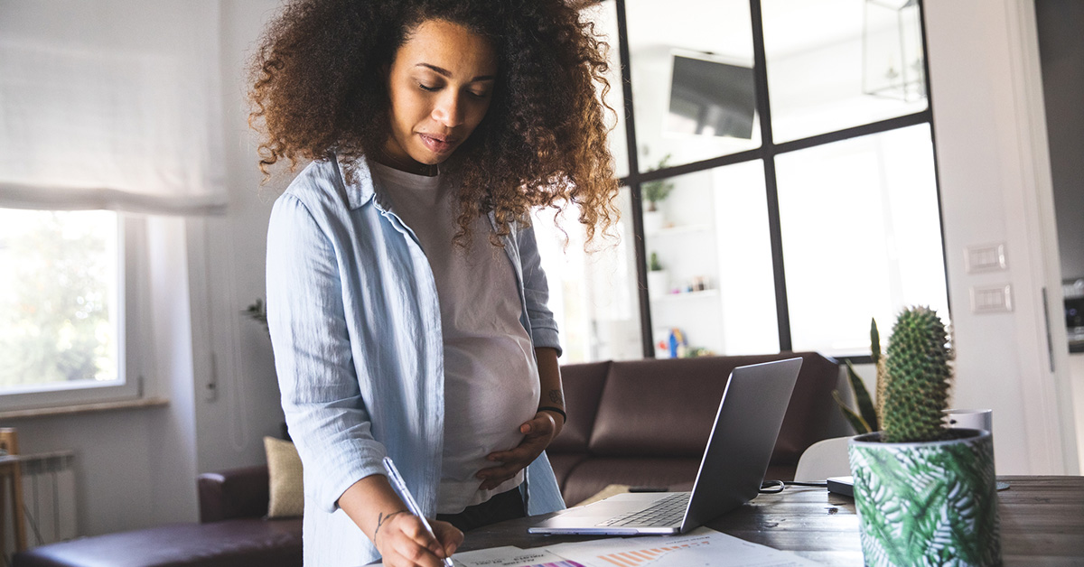 A pregnant Black woman stands at a table writing on a piece of paper.