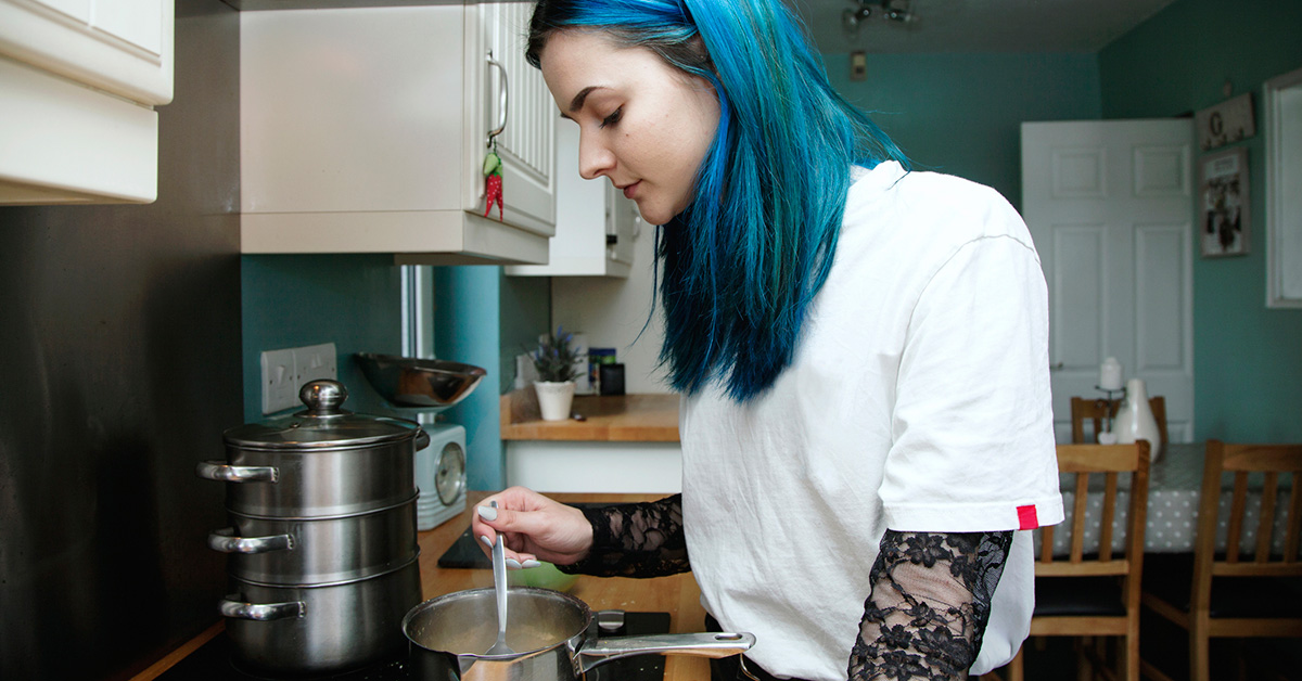 A teen girl with blue hair heats food in a pot over the stove.