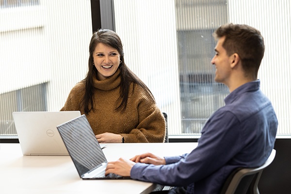 two students smiling at a desk with their laptops