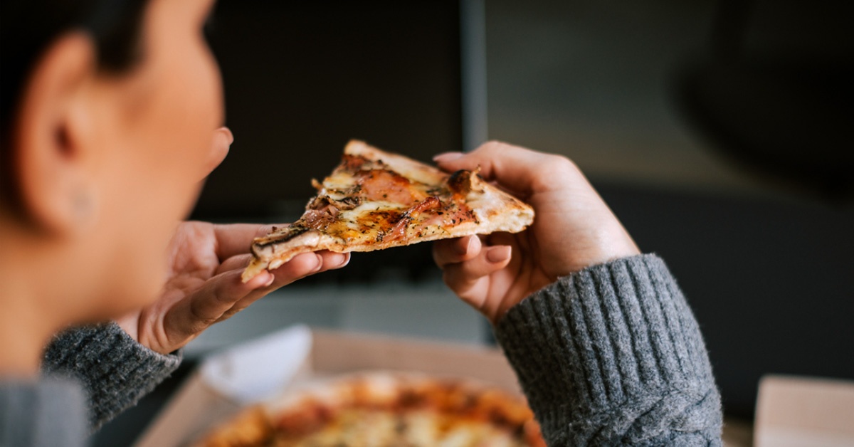 Person holding a slice of pizza up toward their mouth, with rest of the pizza on a table in the background.