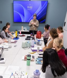 a teacher at a digital board talks to to students at a conference table.