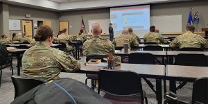 People in military camouflage clothing sit in a classroom facing a man teaching a class. 