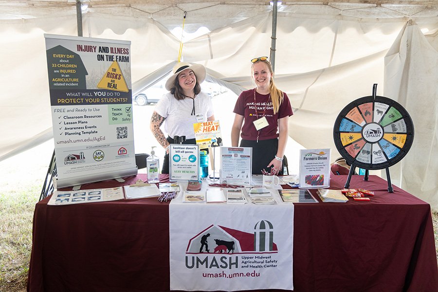 two people standing at a table at farmfest 2023. table includes a poster, and spinning wheel