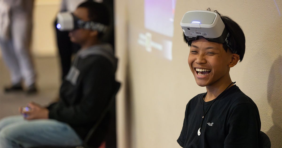 teen wearing VR goggles laughing during drone-flying competition. Other teens are visible in the background