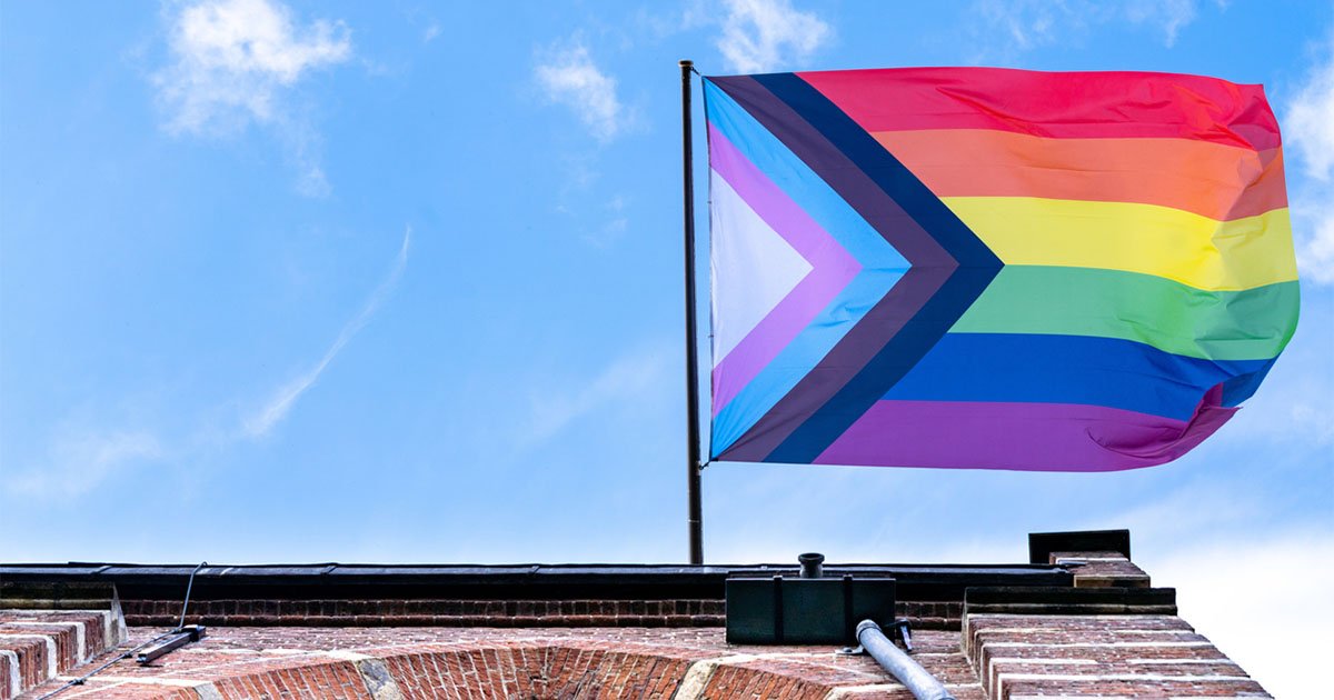 LGBTQ+ flag hangs off a brick building, blue sky in background