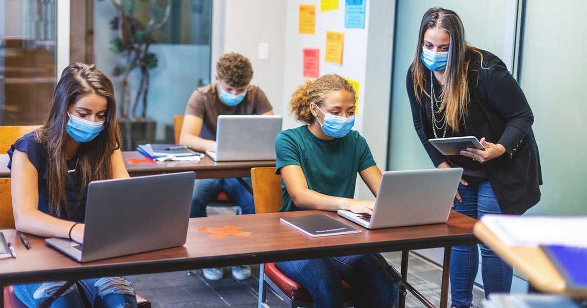 Masked students in a high school classroom working on laptops with a masked teacher standing nearby