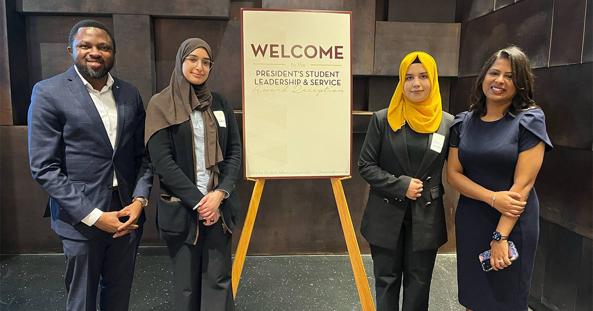 Four School of Public Health students, one man and three women, standing next to a sign that says welcome.