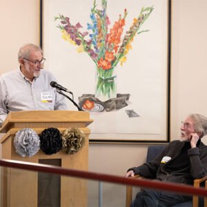 David Jacobs speaking at a lectern with Henry Blackburn seated next to him
