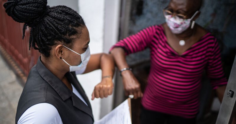 Surveyor greeting senior woman in the doorway - wearing face mask Two Black women wearing masks stand in an apartment hallway. One holds a clipboard and they are bumping elbows.
