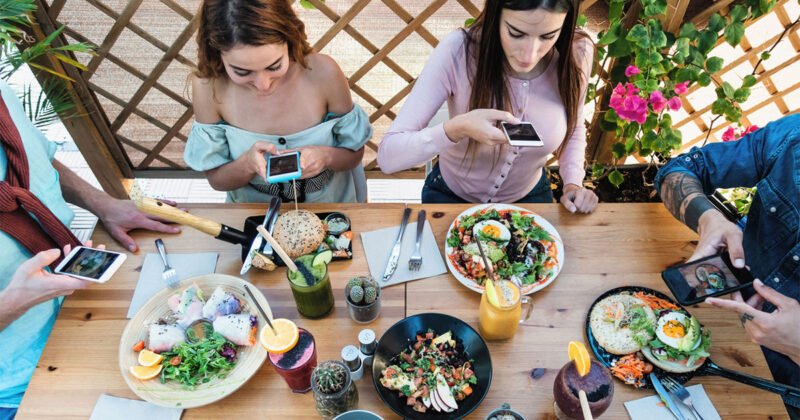 Younger people seated around a table holding phones and taking photos of plates of food