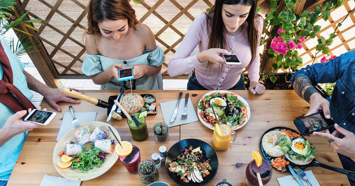 Younger people seated around a table holding phones and taking photos of plates of food