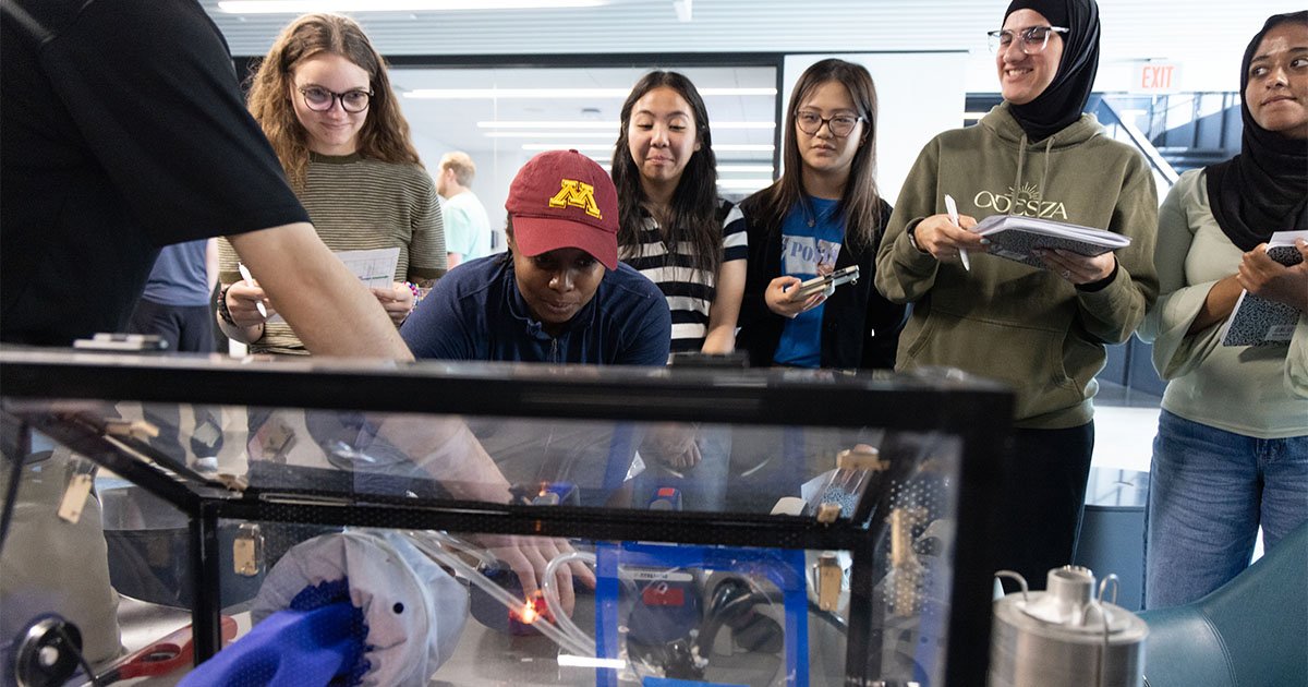 Several students stand around a glass box watching an instructor conduct a science experiment