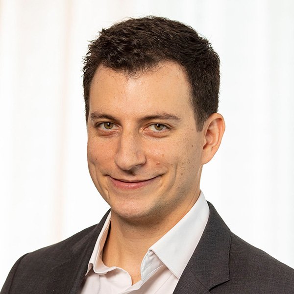 A professional headshot of Eric Jutkowitz with short, dark hair and light eyes, wearing a dark suit jacket over a light-colored collared shirt, smiling. The background is a plain, light color.