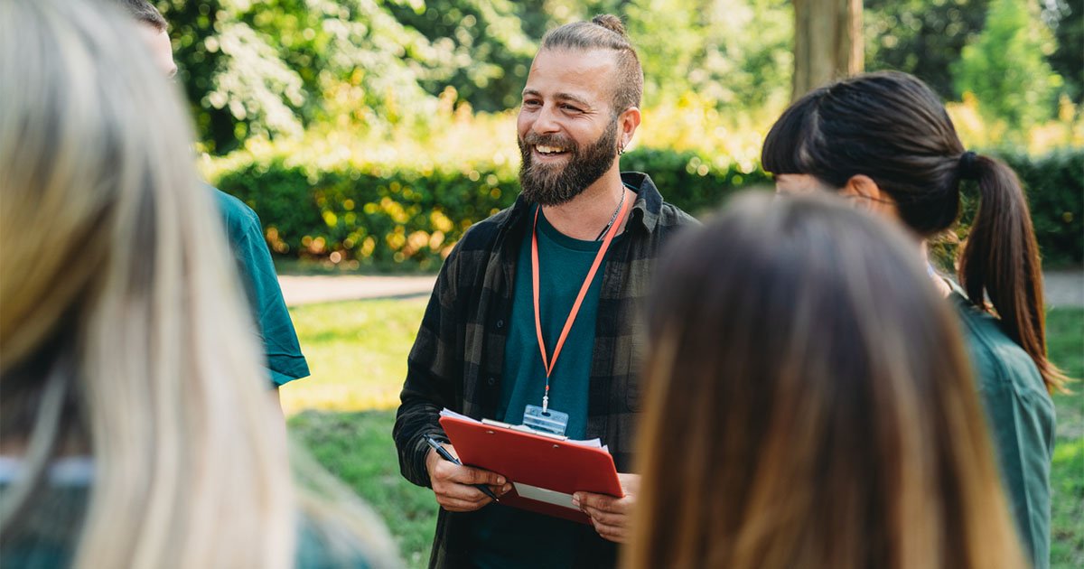 White man holding a clipboard speaking to a group of people standing outside