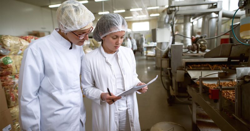 Two people in white coats and hairnets in a food factory doing an inspection